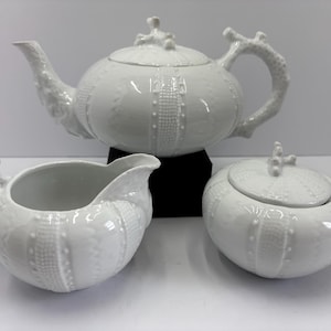 May include: A white ceramic tea set featuring a teapot, creamer, and sugar bowl. The teapot has a curved spout and handle, and the sugar bowl has a lid. The set has a textured, sea urchin-like design.