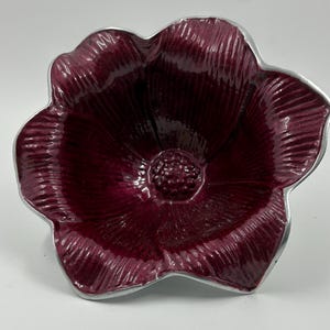 May include: A burgundy-colored, flower-shaped bowl with a silver-colored rim. The bowl has a textured surface and a detailed center, resembling a flower's pistil. The petals are deeply colored, creating a striking contrast with the silver rim.