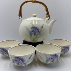 May include: A white ceramic teapot with a woven handle and four matching teacups. Each piece is decorated with a purple iris flower design. The teapot sits on a black base, and the teacups are arranged in front.