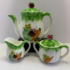 May include: A white ceramic tea set with a vegetable motif. The set includes a teapot, creamer, and sugar bowl. The teapot has a green lettuce-leaf lid with a red radish finial. The creamer and sugar bowl also have green lettuce-leaf accents and vegetable illustrations.