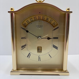 May include: A gold-toned Seiko Quartz clock with a rectangular base and arched top. The clock face features Roman numerals, black hands, and a day display. A gold ring handle sits atop the clock.