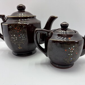 May include: Two brown ceramic teapots with lids, spouts, and handles. The teapots have gold trim and floral designs with red, white, and blue accents. One teapot is larger than the other.