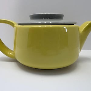 May include: A yellow ceramic teapot with a curved handle and spout. The lid is gray. The teapot is a solid color and sits on a white surface. The teapot is designed for brewing tea.
