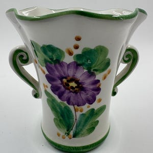 May include: White ceramic vase with a hand-painted purple flower and green leaves. The vase has two handles and a green rim. The flower is the focal point, with yellow dots and green accents.