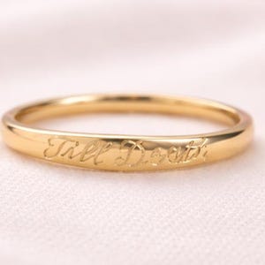 Op de afbeelding: Een goudkleurige ring met de inscriptie "Till Death" gegraveerd op de band. De ring is dun en heeft een glad, gepolijst oppervlak. De ring staat tegen een zachte, roze achtergrond.