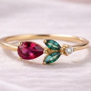 May include: A gold ring with a pear-shaped, deep red gemstone, two green leaf-shaped stones, and a small, round, clear stone. The ring has a delicate, nature-inspired design.