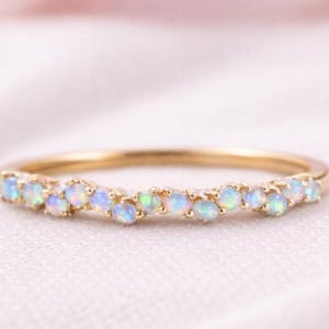 May include: A slender gold ring adorned with a line of small opal gemstones. The opals exhibit a play of colours, including blue, green, and white, set within a polished gold band. The ring is presented on a soft, white fabric background.