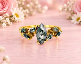 May include: A gold ring featuring a central marquise-cut moss agate stone with a distinct green dendritic inclusion pattern. The band is adorned with smaller marquise-cut moss agate stones arranged in a leaf-like cluster on both sides.