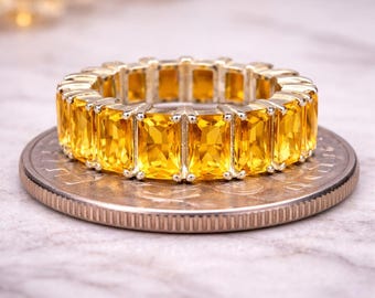May include: A full eternity band ring featuring a continuous row of rectangular, emerald-cut yellow citrine gemstones set in a metallic frame, resting flat on top of a circular United States coin for scale.