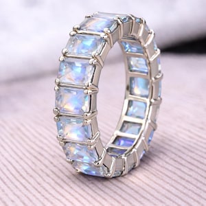 May include: A silver ring featuring a continuous band of rectangular, iridescent gemstones. The gemstones reflect blue, purple, and white. The ring is set on a light pink surface.