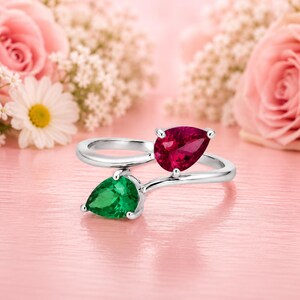 May include: A bypass ring crafted from polished silver-toned metal, featuring two pear-cut gemstones. One stone is a vibrant, deep red, whilst the other is a bright, saturated green. The stones are set in a delicate, open-ended band design.