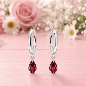 May include: A pair of silver hoop earrings featuring a dangling teardrop-shaped deep red gemstone. The earrings have a polished metallic finish and a secure hinged closure, showcasing a classic design with a vibrant, faceted crimson stone.