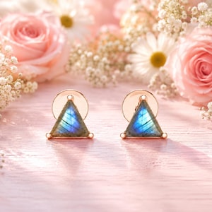 May include: A pair of stud earrings featuring triangular-shaped labradorite gemstones with a blue iridescent flash, set in a metallic frame with circular backings. The stones display a natural grey base color with vibrant blue light reflections.