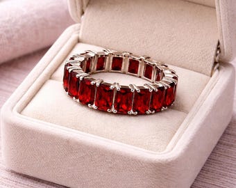 May include: A silver ring featuring a continuous band of rectangular, deep red gemstones. The ring is presented in an open, off-white jewellery box, set against a soft, pinkish backdrop. The gemstones are evenly spaced and reflect light.