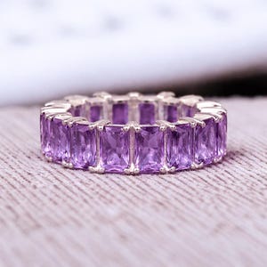 May include: A silver ring featuring a continuous band of rectangular, vibrant purple gemstones. The gemstones are set closely together, creating a full eternity design. The ring is displayed on a textured, neutral-toned surface.