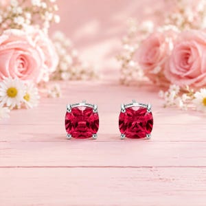 May include: A pair of stud earrings featuring cushion-cut, deep red gemstones set in polished silver-toned metal. Each earring has a four-prong setting that secures the faceted stone, creating a classic and symmetrical appearance for the jewelry.