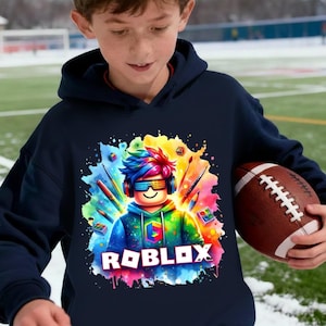 May include: Navy blue hoodie featuring a colorful graphic of a Roblox character with headphones and sunglasses, surrounded by paintbrushes and paint splatters. The word "ROBLOX" is displayed in white block letters. The person is holding a brown football.