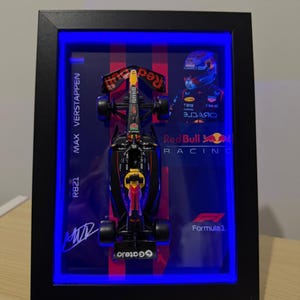 Puede incluir: Coche de modelo de F&oacute;rmula 1 enmarcado, con un dise&ntilde;o en negro, rojo y amarillo. El marco incluye el texto "MAX VERSTAPPEN" y "RB21", junto con el logotipo de Red Bull Racing y la marca Formula 1. El marco tiene un borde negro e iluminaci&oacute;n LED azul.