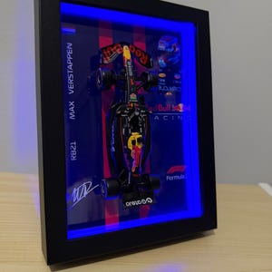 Puede incluir: Una exhibici&oacute;n enmarcada con un modelo de coche de F&oacute;rmula 1 Red Bull Racing RB21. El coche es negro, rojo y amarillo, con logotipos de patrocinadores. El marco tiene un borde negro e iluminaci&oacute;n LED azul. Incluye el nombre "MAX VERSTAPPEN".