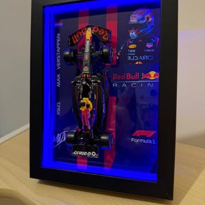 Puede incluir: Un marco tipo vitrina negro que contiene un modelo en miniatura de un coche de F&oacute;rmula 1 de Red Bull Racing. El fondo incluye un retrato de Max Verstappen, el texto Max Verstappen RB21, el logotipo de Red Bull Racing y el logotipo de Formula 1. Una luz LED azul ilumina el interior del marco.
