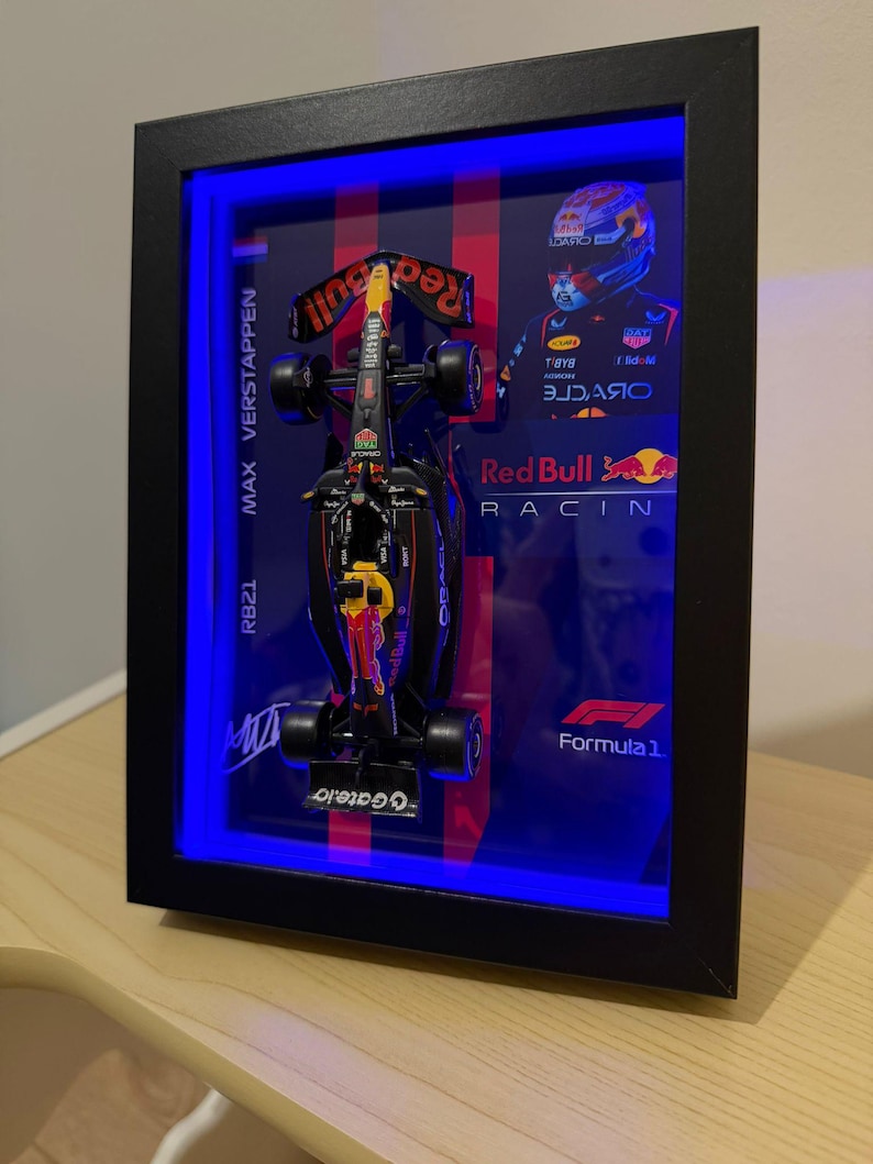 Puede incluir: Un marco tipo vitrina negro que contiene un modelo en miniatura de un coche de F&oacute;rmula 1 de Red Bull Racing. El fondo incluye un retrato de Max Verstappen, el texto Max Verstappen RB21, el logotipo de Red Bull Racing y el logotipo de Formula 1. Una luz LED azul ilumina el interior del marco.