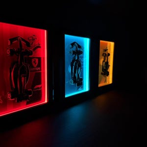 Puede incluir: Tres cajas de luz rectangulares montadas en la pared con siluetas negras de coches de F&oacute;rmula 1. La primera caja brilla en rojo con un logotipo de Ferrari, la segunda en azul y la tercera en amarillo. Cada marco muestra un contorno detallado.