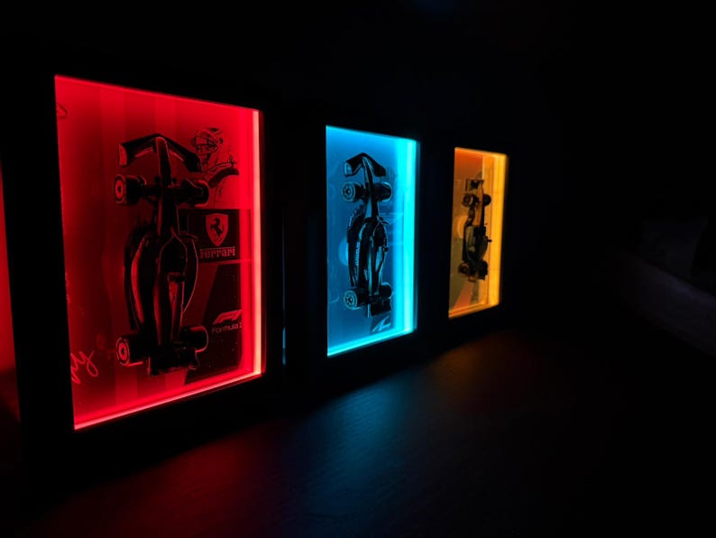Puede incluir: Tres cajas de luz rectangulares montadas en la pared con siluetas negras de coches de F&oacute;rmula 1. La primera caja brilla en rojo con un logotipo de Ferrari, la segunda en azul y la tercera en amarillo. Cada marco muestra un contorno detallado.
