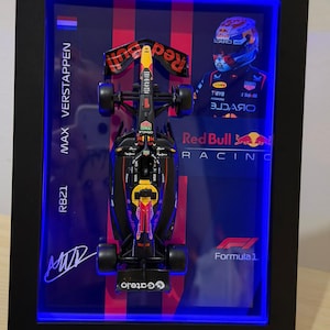 Puede incluir: Un expositor enmarcado con un modelo a escala del coche de F&oacute;rmula 1 Red Bull Racing RB21. El marco negro incluye iluminaci&oacute;n LED azul, una foto de Max Verstappen, el texto Max Verstappen, RB21, Red Bull Racing, Formula 1 y una firma.
