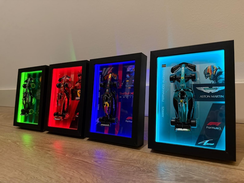 Puede incluir: Cuatro modelos de coches de F&oacute;rmula 1 enmarcados con retroiluminaci&oacute;n LED. Los marcos son negros y los coches se exhiben frente a luces de colores. Un marco presenta un coche Aston Martin con el texto "Fernando Alonso".