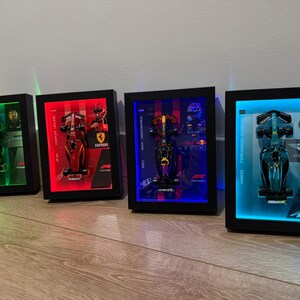 Puede incluir: Cuatro modelos enmarcados de coches de carreras de F&oacute;rmula 1 con retroiluminaci&oacute;n LED. Cada marco tiene un borde negro y presenta un coche diferente con la imagen de un piloto y el logotipo del equipo. Los marcos est&aacute;n iluminados con luces verdes, rojas, azules y cian.