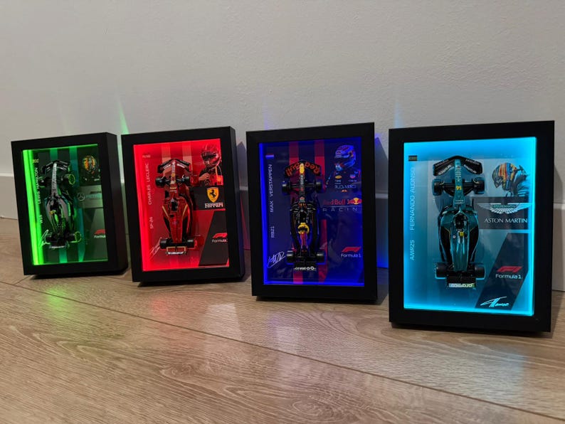 Puede incluir: Cuatro modelos enmarcados de coches de carreras de F&oacute;rmula 1 con retroiluminaci&oacute;n LED. Cada marco tiene un borde negro y presenta un coche diferente con la imagen de un piloto y el logotipo del equipo. Los marcos est&aacute;n iluminados con luces verdes, rojas, azules y cian.