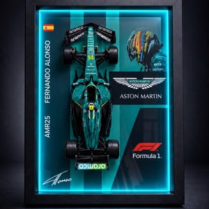 May include: A framed display featuring a scale model of the green Aston Martin AMR25 Formula 1 car driven by Fernando Alonso. The display includes the driver's helmet, the Aston Martin logo, the Formula 1 emblem, and a signature in a black frame.
