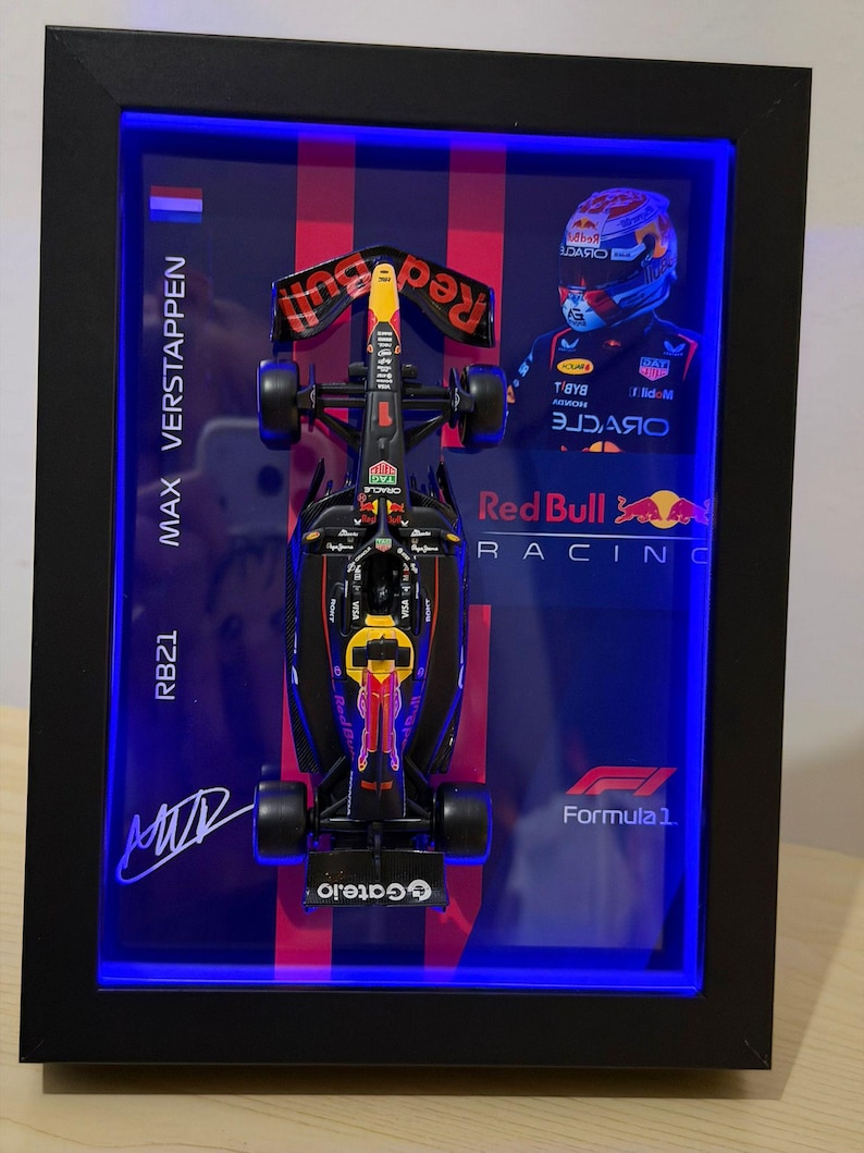 Puede incluir: Un marco decorativo que contiene un coche de F&oacute;rmula 1 Red Bull Racing en miniatura, modelo RB21, pilotado por Max Verstappen. Incluye una foto del piloto, los logotipos de Red Bull Racing y F&oacute;rmula 1, y una firma, todo iluminado con luces LED azules.