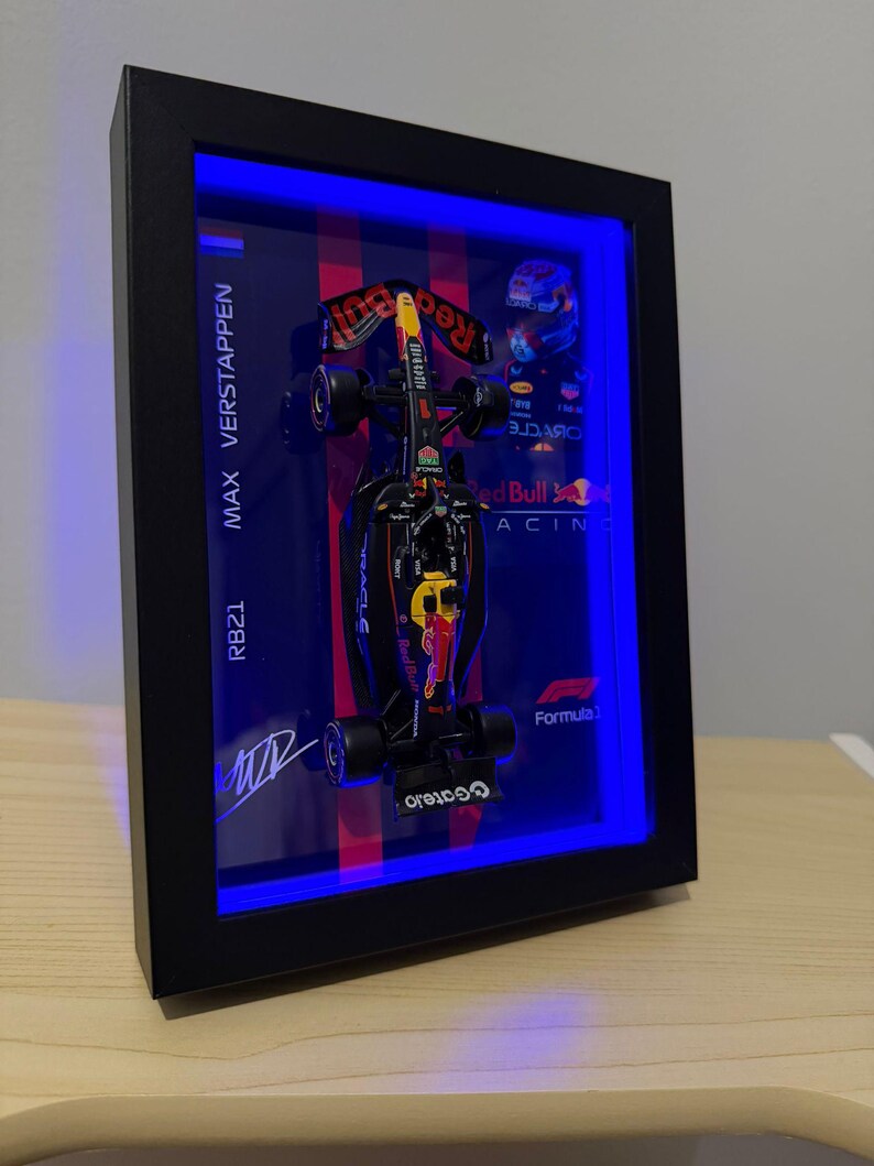 Puede incluir: Un marco de caja negra que contiene un modelo en miniatura del coche de F&oacute;rmula 1 Red Bull Racing RB21. La pantalla cuenta con iluminaci&oacute;n LED azul, el texto MAX VERSTAPPEN, RB21 y Formula 1, una firma impresa y un gr&aacute;fico de un casco.