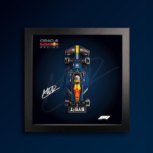 Puede incluir: Una vista superior enmarcada de un modelo de coche de Fórmula 1 de Oracle Red Bull Racing en azul oscuro. La pantalla presenta el logotipo de Oracle Red Bull Racing, una firma blanca y el logotipo de F1 sobre un fondo oscuro. El coche incluye detalles en amarillo y rojo.