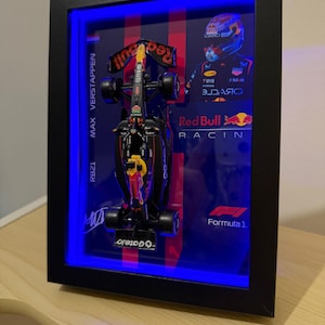 Puede incluir: Modelo enmarcado de un coche de F&oacute;rmula 1 Red Bull Racing. El coche negro y rojo se exhibe en una caja de sombra con iluminaci&oacute;n LED azul. El marco incluye el texto "MAX VERSTAPPEN" y "RB21". Tambi&eacute;n son visibles el logotipo de Red Bull Racing y el logotipo de F&oacute;rmula 1.