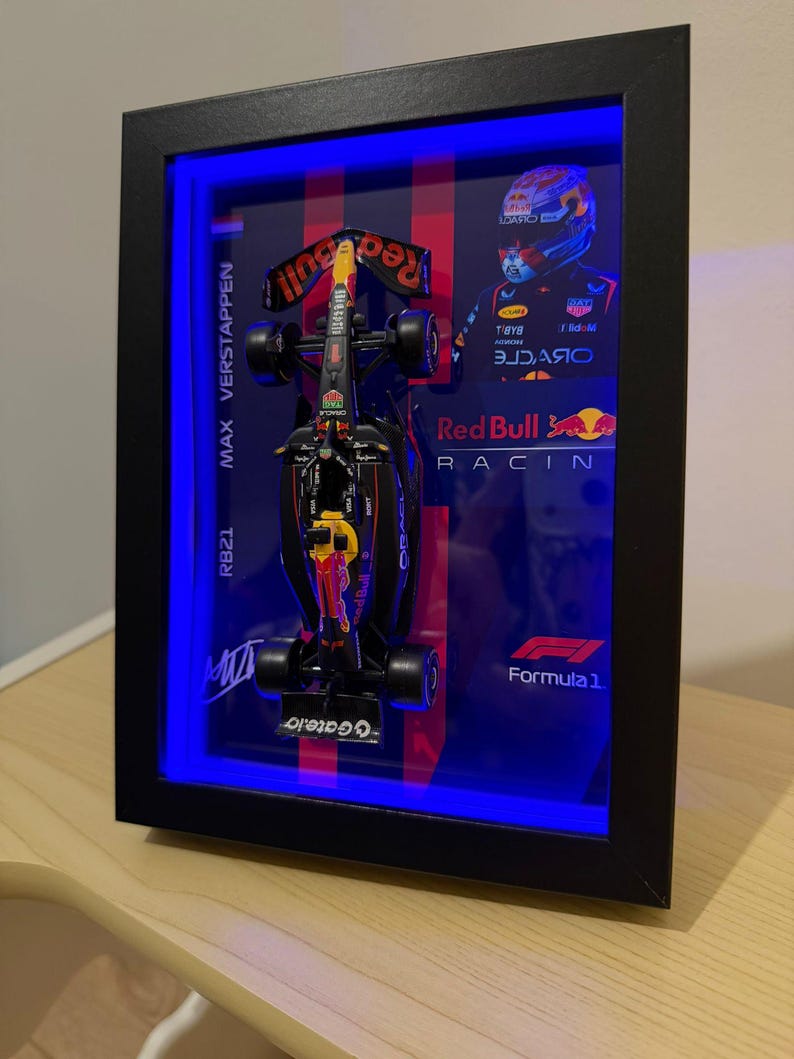Puede incluir: Modelo enmarcado de un coche de F&oacute;rmula 1 Red Bull Racing. El coche negro y rojo se exhibe en una caja de sombra con iluminaci&oacute;n LED azul. El marco incluye el texto "MAX VERSTAPPEN" y "RB21". Tambi&eacute;n son visibles el logotipo de Red Bull Racing y el logotipo de F&oacute;rmula 1.
