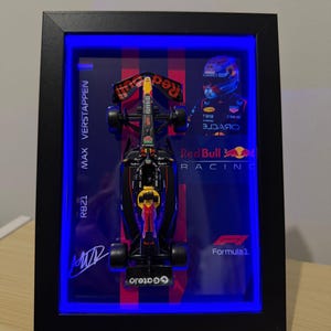 Puede incluir: Un expositor enmarcado con un modelo a escala de un coche de F&oacute;rmula 1 de Red Bull Racing. El fondo incluye el texto Max Verstappen, RB21, Red Bull Racing y Formula 1, junto a una imagen del piloto y una firma. El marco est&aacute; iluminado con luces LED azules.