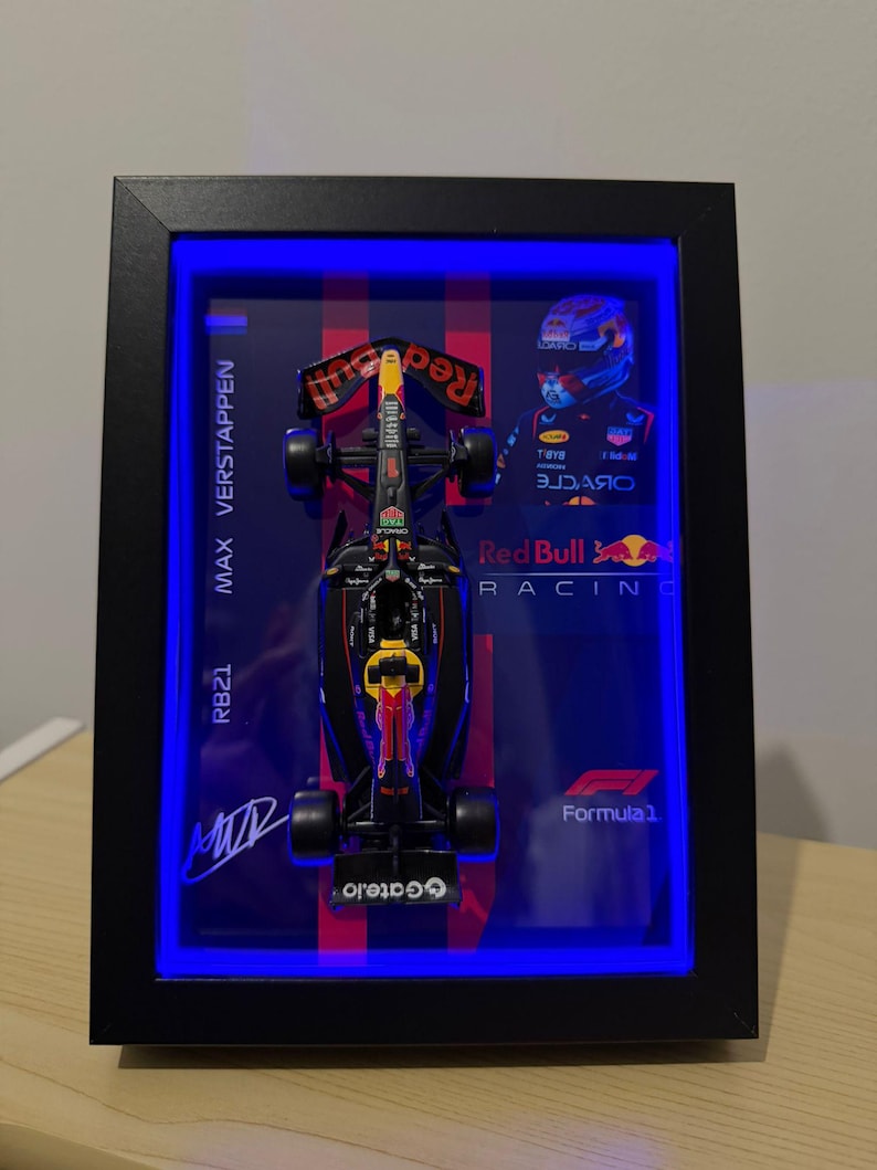 Puede incluir: Un expositor enmarcado con un modelo a escala de un coche de F&oacute;rmula 1 de Red Bull Racing. El fondo incluye el texto Max Verstappen, RB21, Red Bull Racing y Formula 1, junto a una imagen del piloto y una firma. El marco est&aacute; iluminado con luces LED azules.