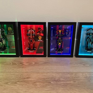 Puede incluir: Cuatro marcos negros rectangulares tipo vitrina que contienen coches de carreras de F&oacute;rmula 1 en miniatura. Cada marco cuenta con retroiluminaci&oacute;n LED en verde, rojo, azul o cian, con nombres de pilotos, logotipos y gr&aacute;ficos impresos.