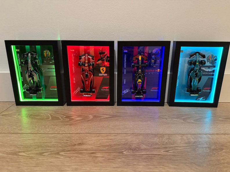 Puede incluir: Cuatro marcos negros rectangulares tipo vitrina que contienen coches de carreras de F&oacute;rmula 1 en miniatura. Cada marco cuenta con retroiluminaci&oacute;n LED en verde, rojo, azul o cian, con nombres de pilotos, logotipos y gr&aacute;ficos impresos.