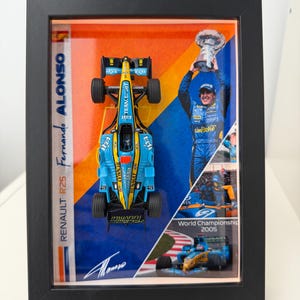 Puede incluir: Exhibición enmarcada con un modelo de coche de carreras Renault R25 azul y amarillo. La exhibición incluye imágenes de Fernando Alonso y el texto "Fernando Alonso" y "Campeonato del Mundo 2005". El marco es negro.