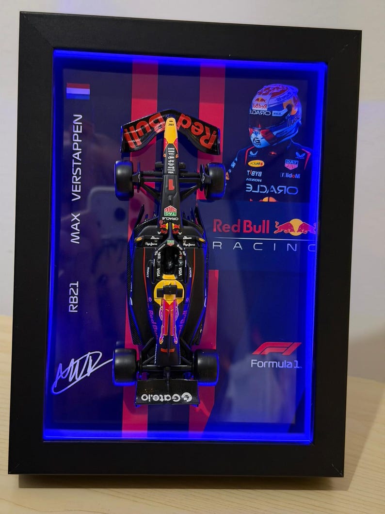 Puede incluir: Una exhibici&oacute;n enmarcada con un modelo de coche de F&oacute;rmula 1 Red Bull Racing RB21. El coche negro, rojo y amarillo est&aacute; posicionado en el centro, con el nombre y la firma del piloto Max Verstappen. El marco tiene una luz LED azul.