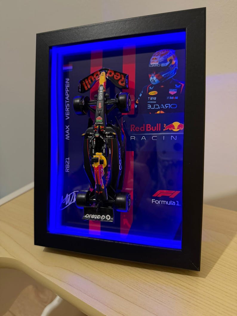 Puede incluir: Un marco tipo vitrina negro que contiene un modelo a escala de un coche de carreras de F&oacute;rmula 1, un retrato de Max Verstappen con casco y el logotipo de Red Bull Racing. La vitrina est&aacute; iluminada con luces LED azules e incluye texto.