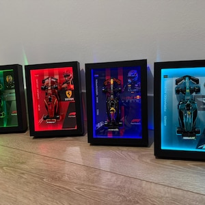 Puede incluir: Cuatro marcos tipo caja negros rectangulares que contienen modelos de coches de carreras de F&oacute;rmula 1. Cada marco cuenta con retroiluminaci&oacute;n LED en verde, rojo, azul o azul claro, con nombres de pilotos, logotipos de equipos y marcas F1.