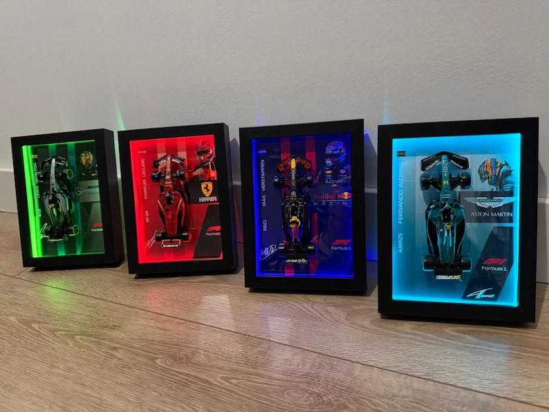 Puede incluir: Cuatro marcos tipo caja negros rectangulares que contienen modelos de coches de carreras de F&oacute;rmula 1. Cada marco cuenta con retroiluminaci&oacute;n LED en verde, rojo, azul o azul claro, con nombres de pilotos, logotipos de equipos y marcas F1.