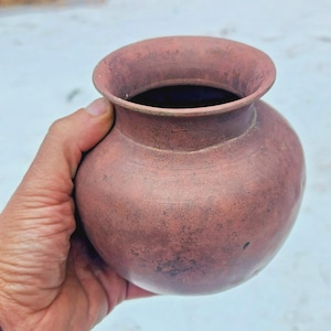 May include: A small, round, terracotta-colored pot with a wide opening. The pot has a slightly textured surface and a dark interior. The pot is held in a hand against a blurred, snowy background.