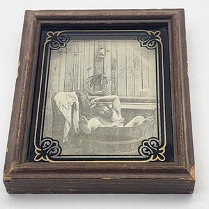 May include: A framed black and white print depicting a person bathing in a wooden tub. The print is set within a dark brown wooden frame with decorative corner accents. The image shows a vintage scene.