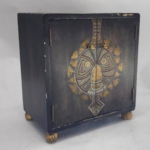 May include: A dark, square cabinet with a decorative face design on the front. The face is made of gold and multi-colored beads. The cabinet has two doors and sits on four gold ball feet.