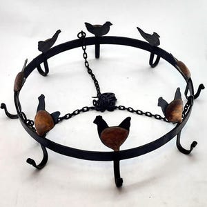 May include: A black metal decorative trivet with a circular design. The trivet features seven stylized chicken figures with brown and black details. A chain connects the center to the ring, and the trivet has curved feet.